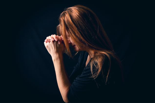 Imagem de uma mulher ruiva usando uma blusa preta. Ela está ajoelhada fazendo a sua oração.