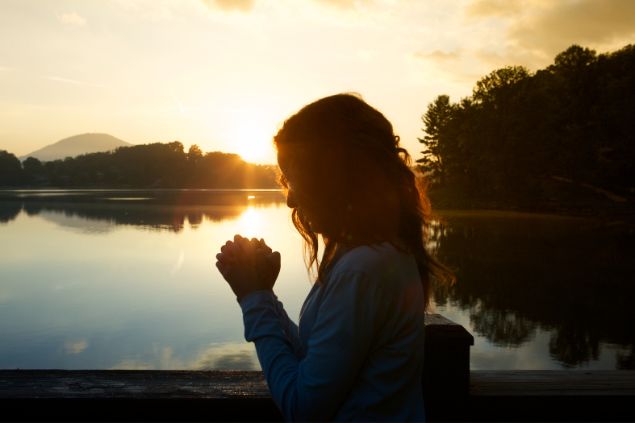 Jjneff / Getty Images Signature / Canva Imagem de uma mulher em pé, fazendo a sua oração e ao fundo um lindo pôr do Sol em frente a um lago.
