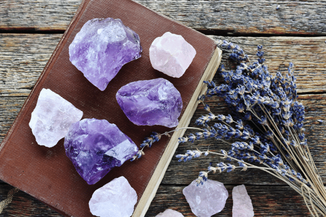 Imagem de várias pedras ametistas distrubuídas sobre um livro de capa marrom sobre uma mesa de madeira. Ao lado, algumas pedras de quartzo rosa e um ramo de flor de lavanda.