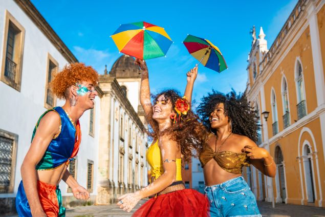 Imagem com 3 pessoas, duas mulheres e um homem, fantasiadas, pulando o carnaval em uma rua do Pelourinho.