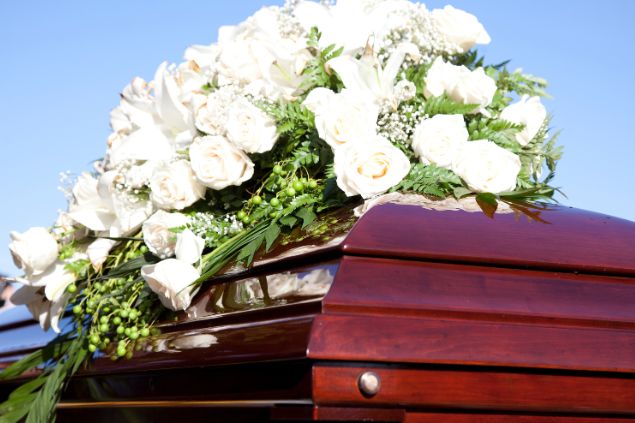 Imagem de um caixão fechado e sobre ele, um arranjo de rosas brancas, simbolizando a morte de uma pessoa conhecida.