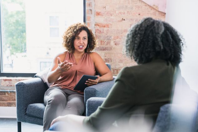 Imagem de duas pessoas sentadas em um sofá em uma sala de estar. Elas estão trocando conselhos, representando o signo de peixes.