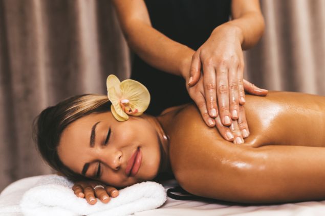 Imagem de uma mulher deitada sobre uma toalha em cima de uma maca, recebendo uma massagem revitalizante em suas costas.