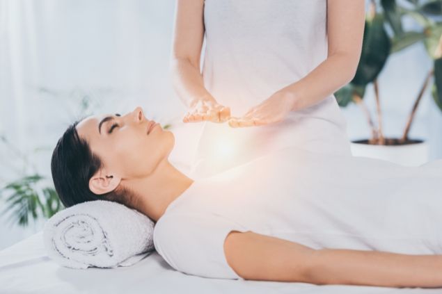 Imagem de uma mulher vestida de roupa branca, deitada em uma maca, recebendo o reiki das mãos de uma terapeuta.