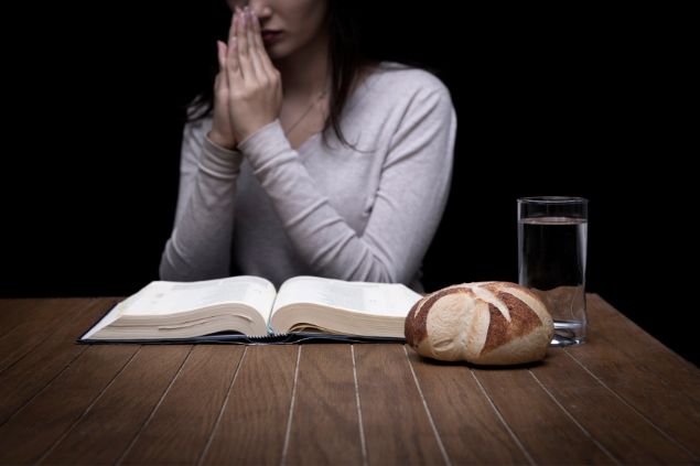 Imagem de uma jovem sentada. Sobre uma mesa a bíblia aberta, um copo com água e um pão, simbolizando o jejum durante o período da Quaresma.