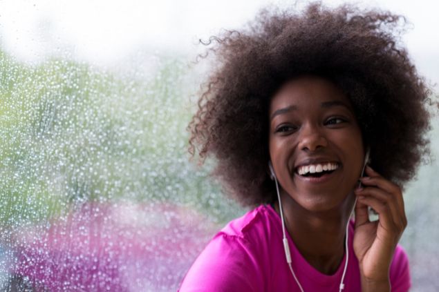 Imagem de uma mulher preta, usando uma camiseta roxa e um fone de ouvido. Ela está feliz e sua imagem simboliza o Dia Internacional da Mulher.