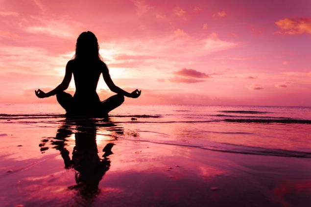 Imagem de uma praia e um lindo pôr do Sol avermelhado. Em destaque, uma mulher sentada na areia, próximo à água fazendo a sua meditação.