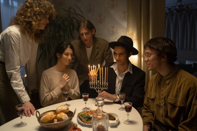 Imagem de uma família judia reunida em uma sala de jantar. Em destaque, um homem acendendo velas no castiçal, celebrando a sexta-feira santa na religião deles.