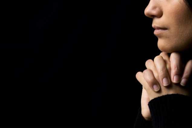 Imagem de fundo preto e em destaque o rosto de uma mulher com as mãos unidas em sinal de oração.