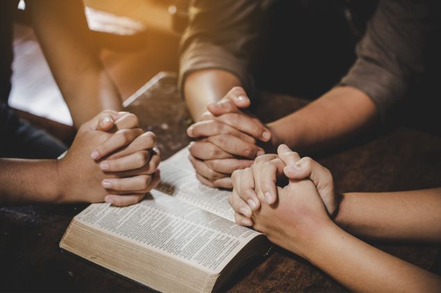 Imagem de um grupo de pessoas em oração. Eles estão sentados e sobre uma mesa, uma bíblia aberta e suas mãos estão em cima dela.