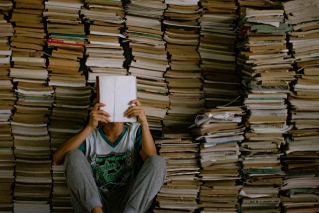 Imagem de uma pessoa do signo de leão, sentado no chão, fazendo uma leitura em um livro. Ao fundo, uma estante cheia de livros.
