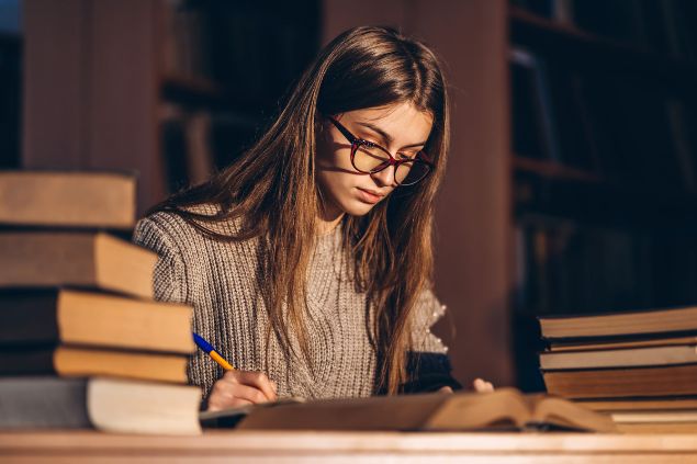 Imagem de uma mulher do signo de escorpião estudando em uma biblioteca. Sobre a mesa, vários outros livros de estudos.