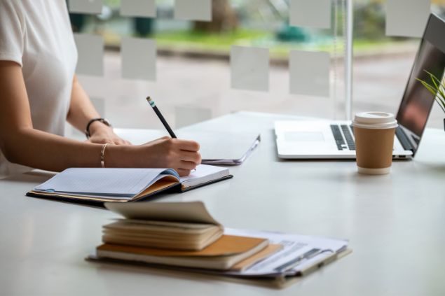 Imagem de uma mulher do signo de capricórnio sentada na frente do computador. Ela está estudando e fazendo várias anotações em um caderno à parte.