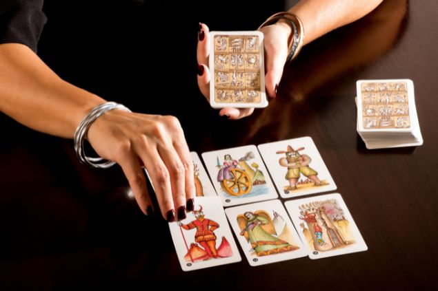 Imagem das mãos de uma mulher cartomante, tirando as cartas do baralho dispostas sobre uma mesa coberta com uma toalha na cor preta.
