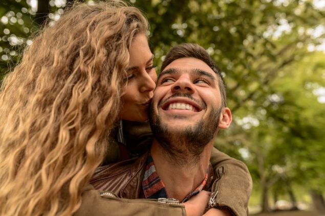 Imagem de um casal. A mulher está dando um beijo no rosto do seu parceiro. Ambos riem e trazem o conceito de felicidade na relação.