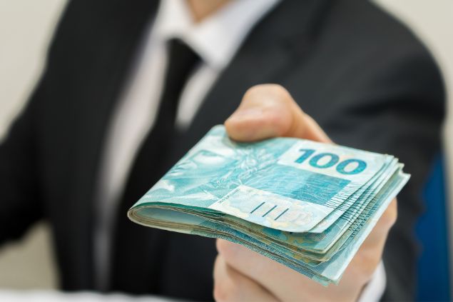 Imagem de um homem executivo, usando um terno, segurando um bolo de dinheiro em notas de cem reais.