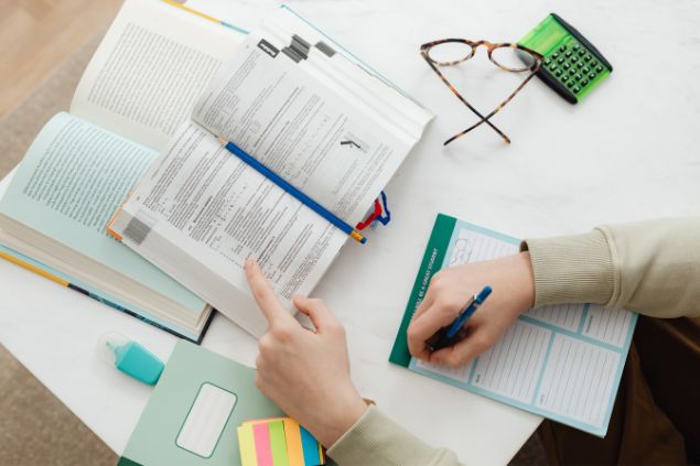 Imagem de um professor universitário estudando e preparando a sua aula semanal. Sobre a mesa, vários post-its, óculos de grau, borracha e caneta.