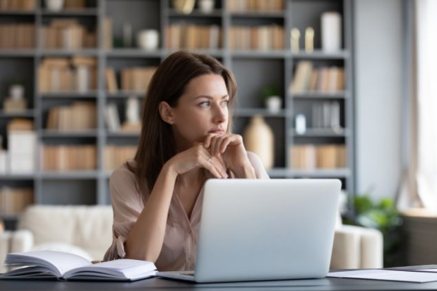 Imagem de uma mulher designer. Ela está de frente com um computador, olhando pela janela e pensando no conceito criativo de suas peças.