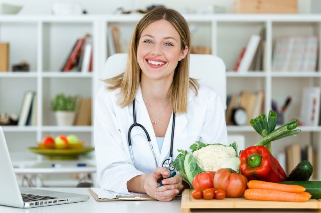 Imagem de uma nutricionista em sua sala de atendimento. Sobre a sua mesa, vários tipos de vegetais.