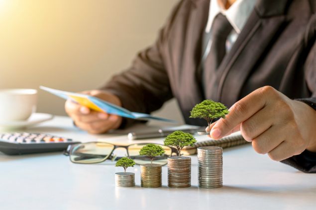 Imagem de um homem que trabalha no departamento financeiro de uma empresa. Sobre sua mesa, moedas e muitos papéis.