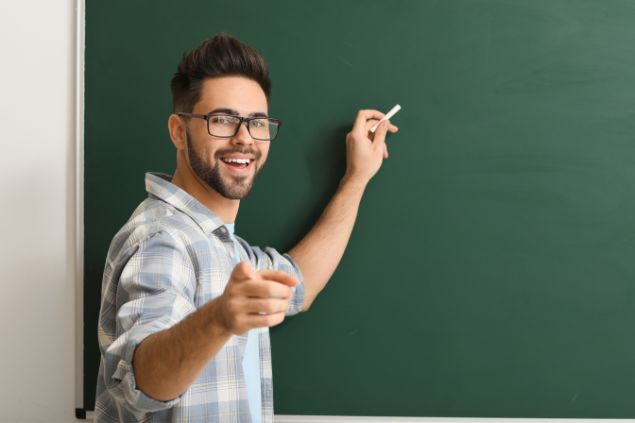 Imagem de um jovem professor segurando um giz branco para escrever na lousa verde.