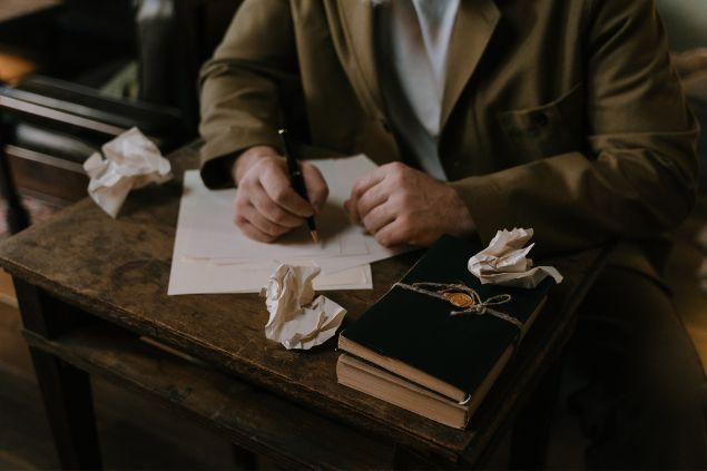 Imagem de um escritor. Sobre a sua mesa, vários papéis amassados e um livro. Ele está escrevendo em um bloco de folhas soltas.