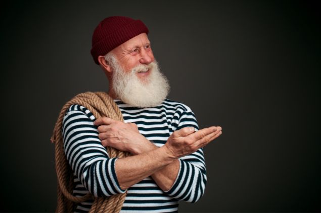 Imagem de um senhor marinheiro usando uma blusa listrada e uma touca de lã sobre a cabeça. Ele também segura uma corda em seu ombro.