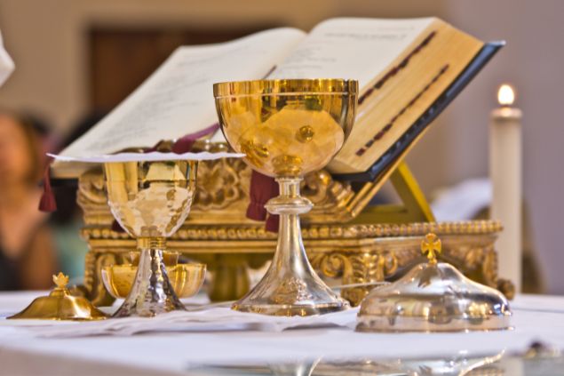 Imagem de um altar da igreja católica durante a celebração da missa. Nele constam: dois cálises, uma vela, uma bíblia e um pequeno sino.