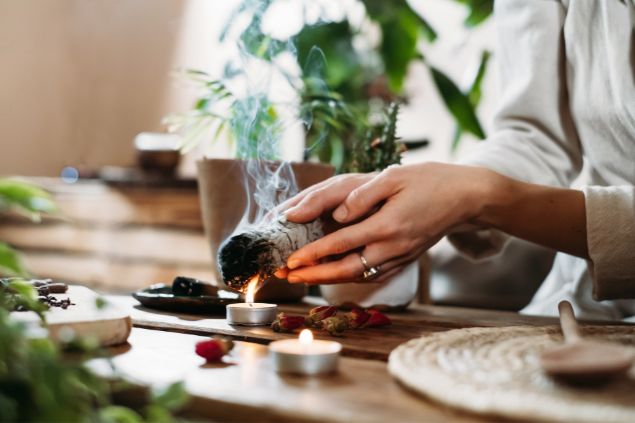 Imagem de uma mulher preparando um ritual de limpeza enérgética. Ela usa uma mocha, uma vela, uma colher de pau e alguns elementos de madeira.