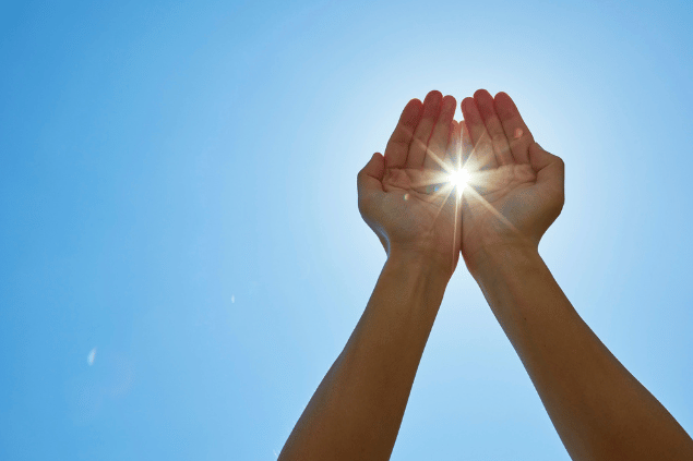 Uma pessoa está com os braços levantados e as mãos juntas em frente da luz do sol. Ao fundo, há o céu azul.