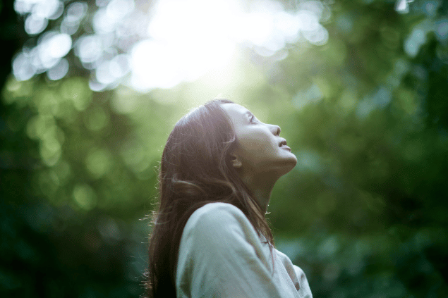 Uma mulher está em uma floresta. Ela levanta a cabeça e sente os raios de sol na pele.
