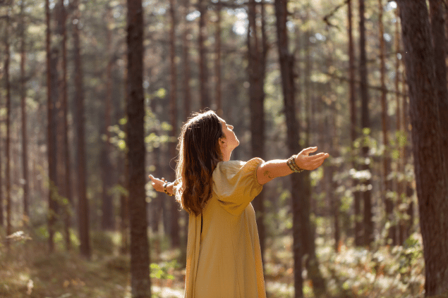 Syda Productions / Canva Uma mulher está com os braços abertos em uma floresta. Ela levanta a cabeça, fecha os olhos e sente os raios de sol na pele.