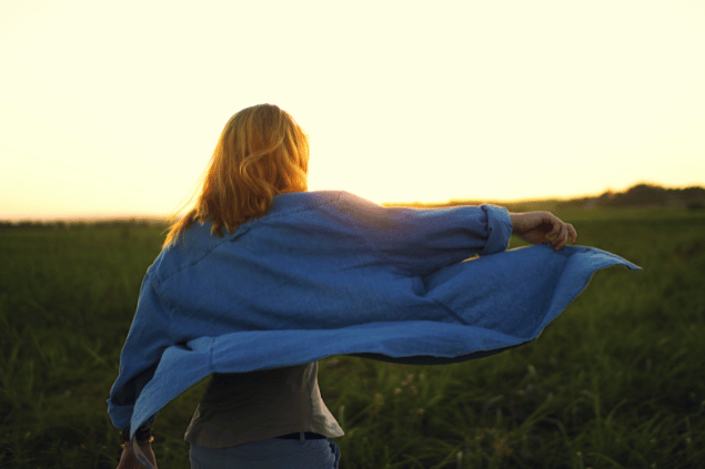 Uma mulher está com os braços abertos em um ambiente de natureza. Ela segura sua camisa esvoaçante e sente os raios de sol na pele.