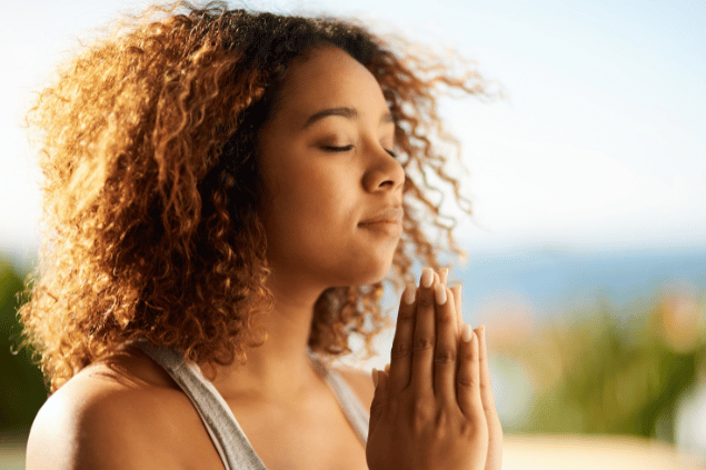 Peopleimages / Getty Images Signature / Canva Uma mulher está com as mãos juntas em forma de oração. Ela medita e sente os raios de sol na pele.