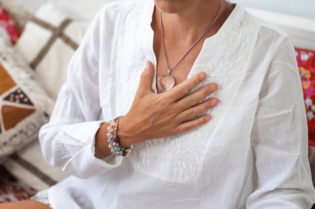 Uma mulher está com a mão no peito em sinal de paz, meditação. Ela usa roupa branca e um colar de cristal.