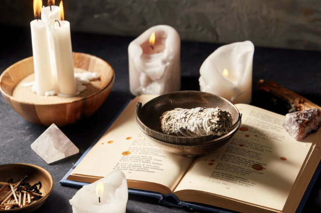 Na imagem, há alguns recipientes com sálvia branca, velas e fósforos em cima de uma mesa. Além disso, há cristais e um livro aberto.