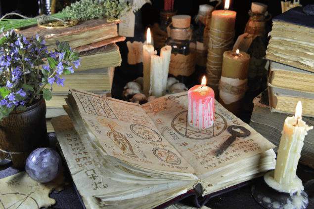Na imagem, há um livro aberto, velas acesas, livros fechados, um cristal e flores em cima de uma mesa.