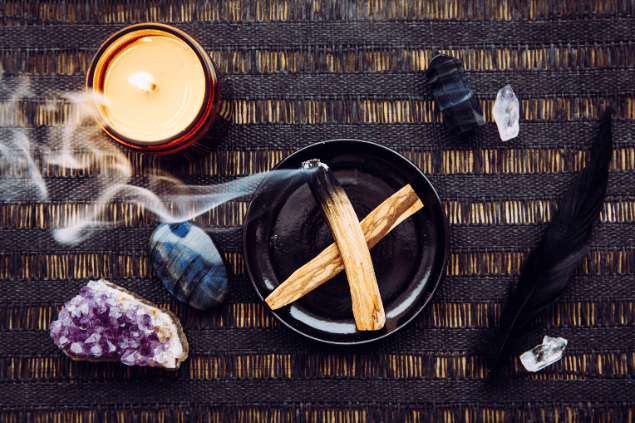 Na imagem, há um recipiente com palo santo em cima de uma mesa. Há uma vela, pedras e uma pena.