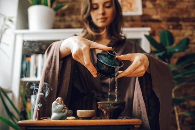 Na imagem, as mãos de uma mulher enchendo um recipiente com água estão em destaque. Há um incenso, uma estátua e uma pedra em cima de uma mesa.