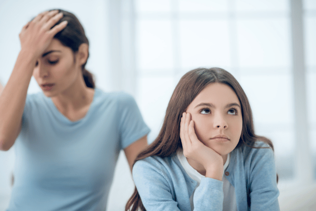 Mulher com a mão na cabeça enquanto sua filha está virada de costas apoiando a cabeça em sua mão direita