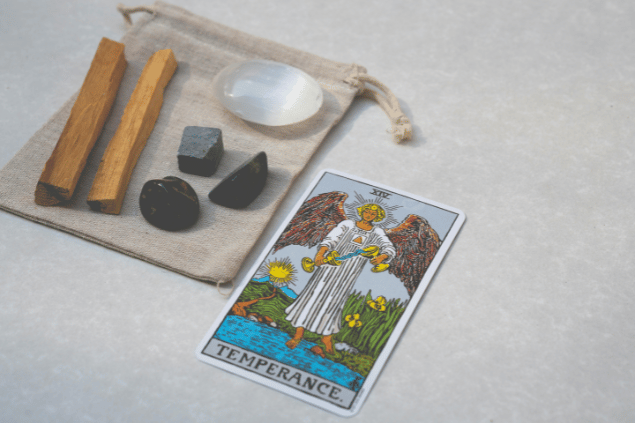 A carta A Temperança do Tarot está em destaque na imagem. Ela está em cima de uma mesa e, ao lado, há cristais.