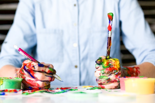 Homem branco com mãos sujas de tintas coloridas, enquanto segura dois pincéis.