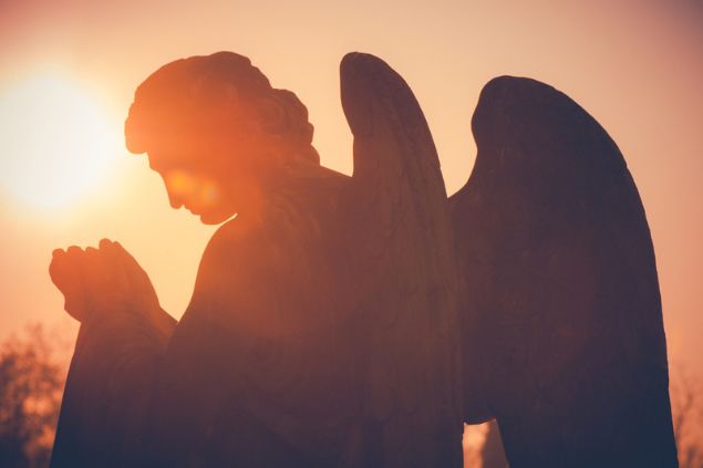 Silhueta de estátua de um anjo orando. O Sol o ilumina.