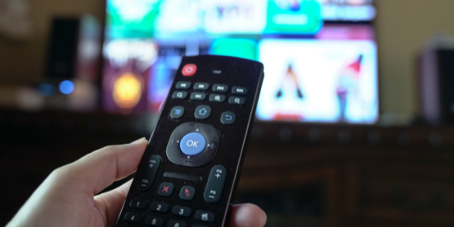 Pessoa segura controle remoto e o aponta para televisão ligada.