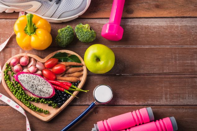 Prato de madeira no formato de coração, preenchido com vários alimentos saudáveis. Ao redor, outros alimentos, uma fita métrica, parte de um tênis, um haltere e parte de um estetoscópio.
