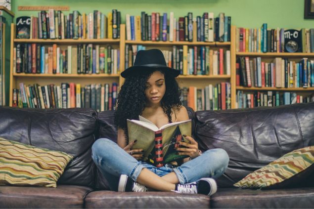 Mulher lendo um livro sentada em um sofá, com uma grande estante de livros atrás.