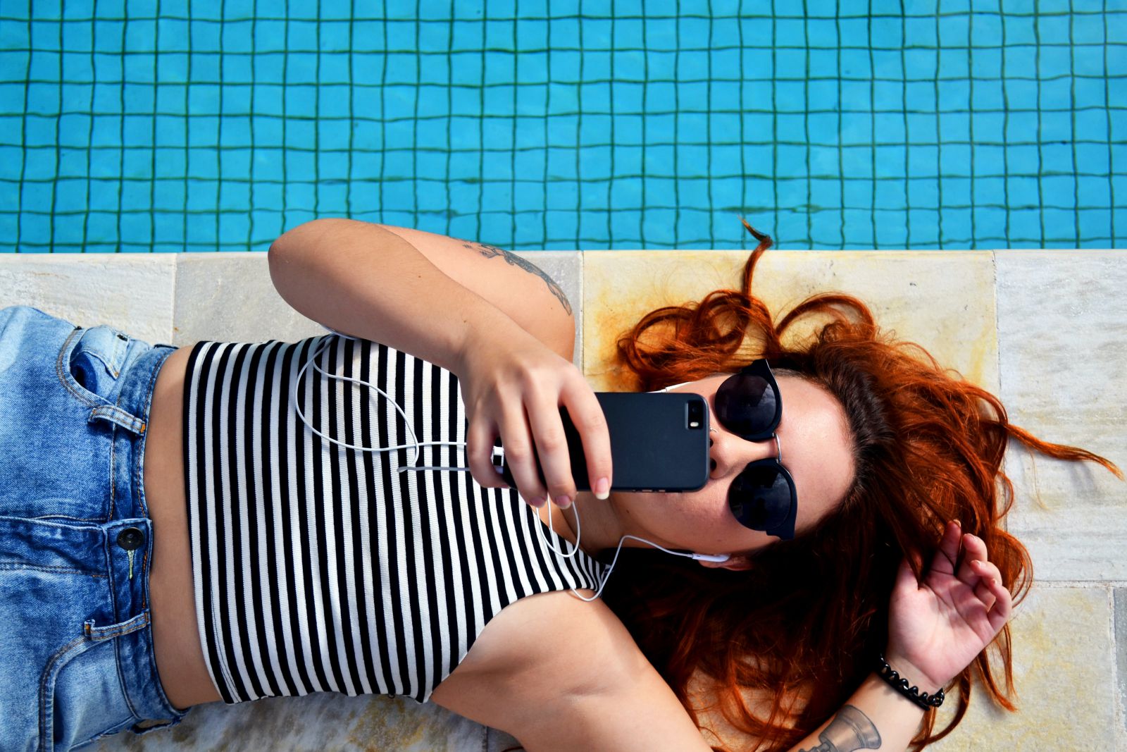 Mulher deitada ao lado da piscina segurando celular e ouvindo música pelo fone de ouvido