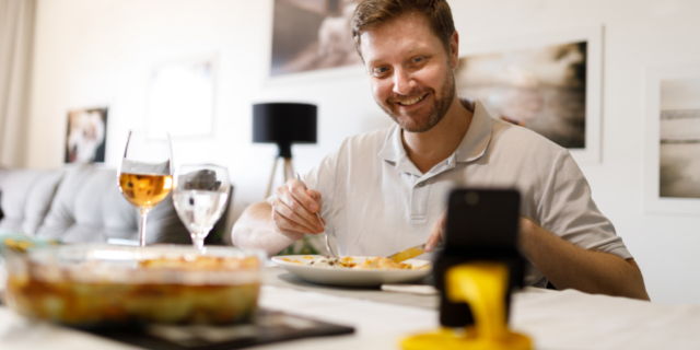 Homem sentado à mesa tem uma refeição em frente do celular que está sobre a mesa. Ele sorri.