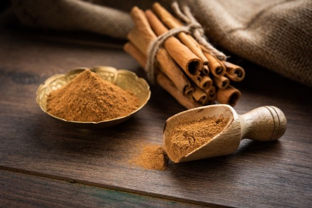Pequeno recipiente com canela em pó. Ao lado, cascas de canela amarradas e uma colher medidora com uma dose de canela.