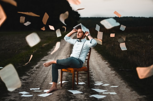 Homem branco sentado numa cadeira no meio da estrada, segurando livro na cabeça, e folhas voando ao redor.